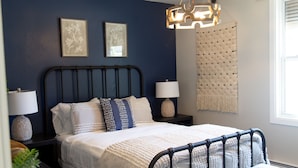 Un dormitorio con cama, dos mesitas de noche, una lámpara colgante y una ventana con persianas.