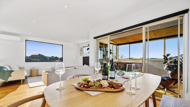 Un comedor moderno con una mesa de madera dispuesta para una comida, un amplio ventanal con vistas y una zona de estar acogedora con un sofá y sillones.