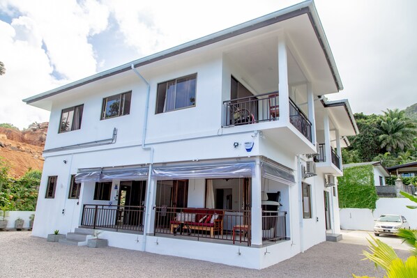 A two-story white building with a balcony, a covered outdoor seating area, and a car parked in the driveway.