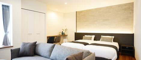A modern hotel room with a grey sofa, a double bed with white bedding, a wooden headboard, and a desk with a chair.