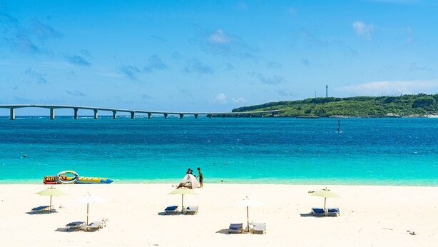 Ein Strand mit weißem Sand, klarem türkisblauem Wasser und einer Brücke im Hintergrund. Liegestühle und Sonnenschirme sind vorhanden.