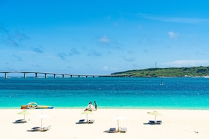 Ein Strand mit weißem Sand, klarem türkisblauem Wasser und einer Brücke im Hintergrund. Liegestühle und Sonnenschirme sind vorhanden.
