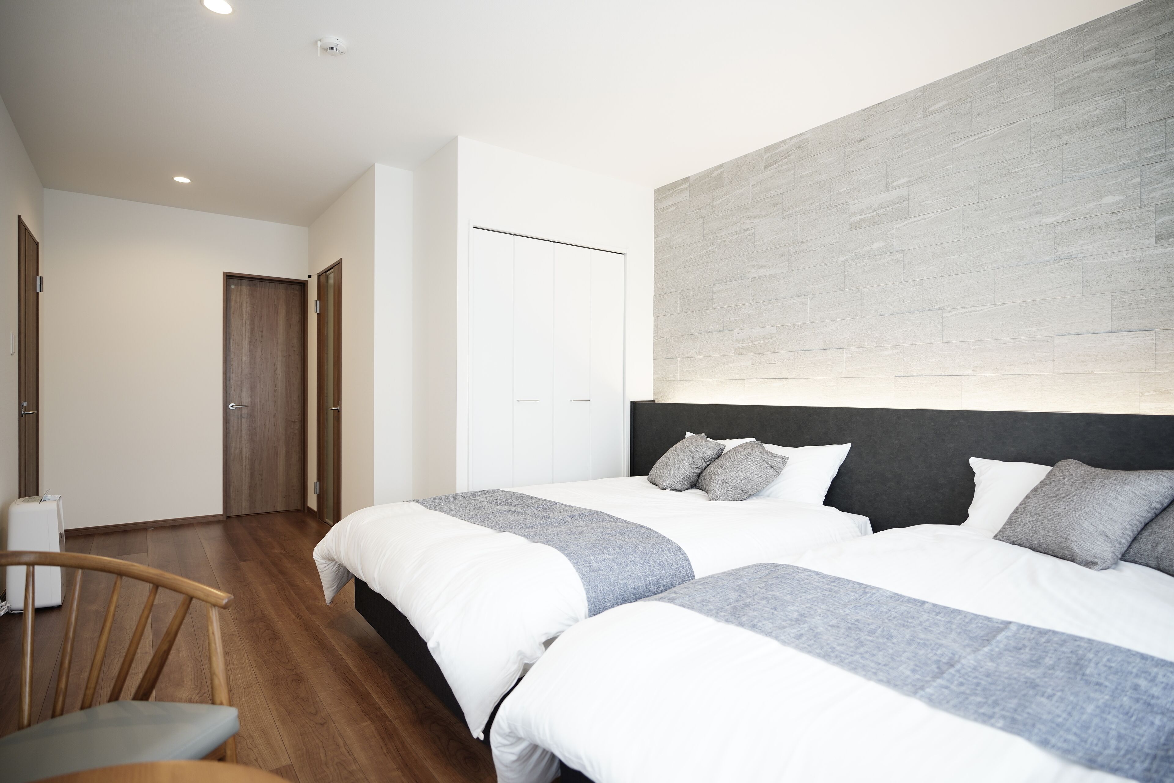 A modern hotel room with two beds, a wooden chair, a white wardrobe, and a grey accent wall.