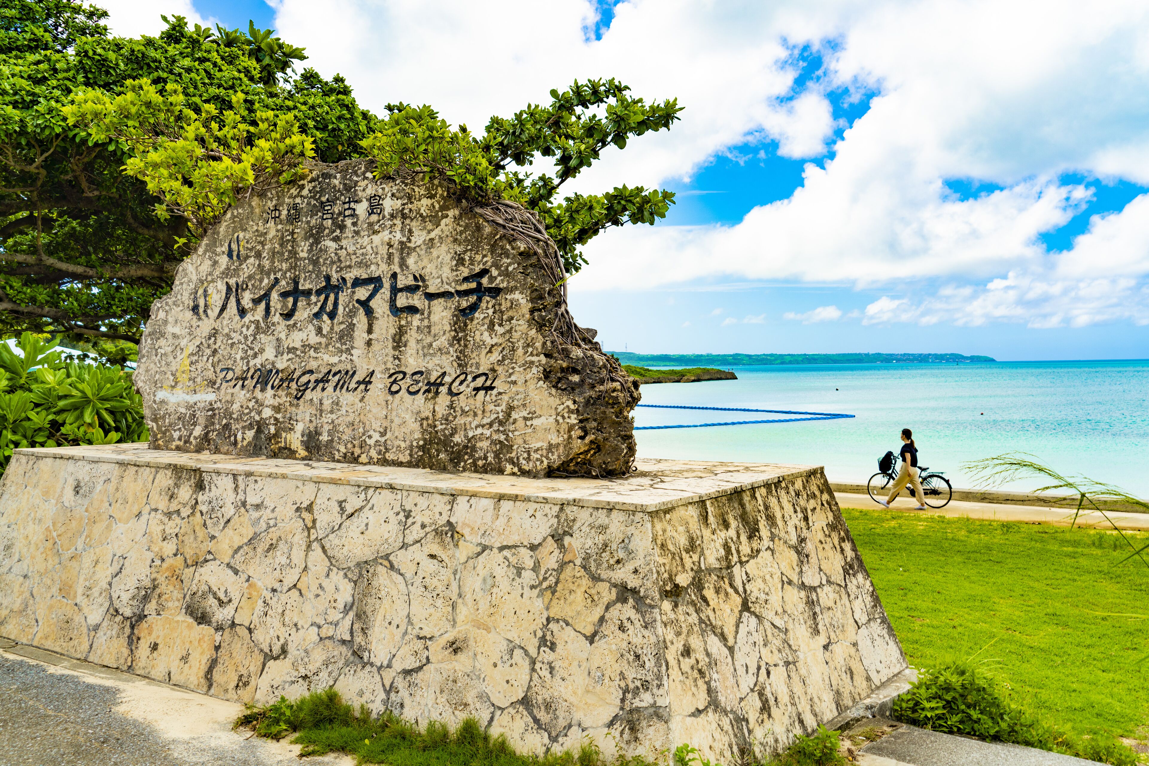 A stone monument with inscriptions, a clear view of the sea, and a cyclist on a path.