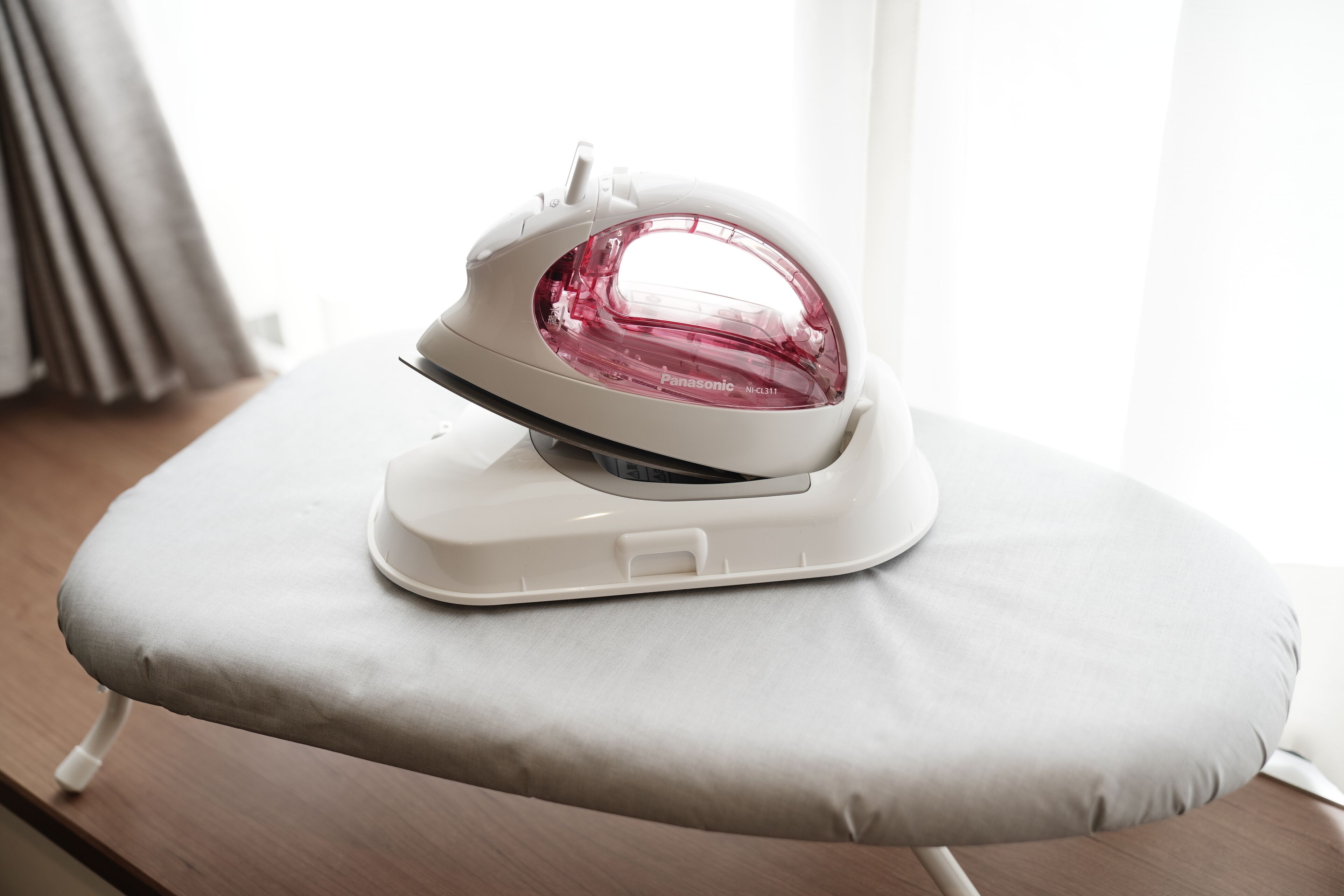 A white iron on a cushioned ironing board in a room with a wooden floor and a window with curtains.