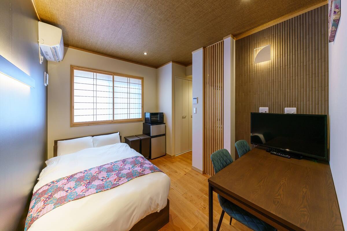 A hotel room with a bed, a desk with a computer, a TV, and a window with blinds.