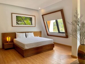 Une chambre avec un lit en bois, une table de chevet avec une lampe, un tableau encadré au mur, une fenêtre donnant sur la verdure et un vase avec des branches.