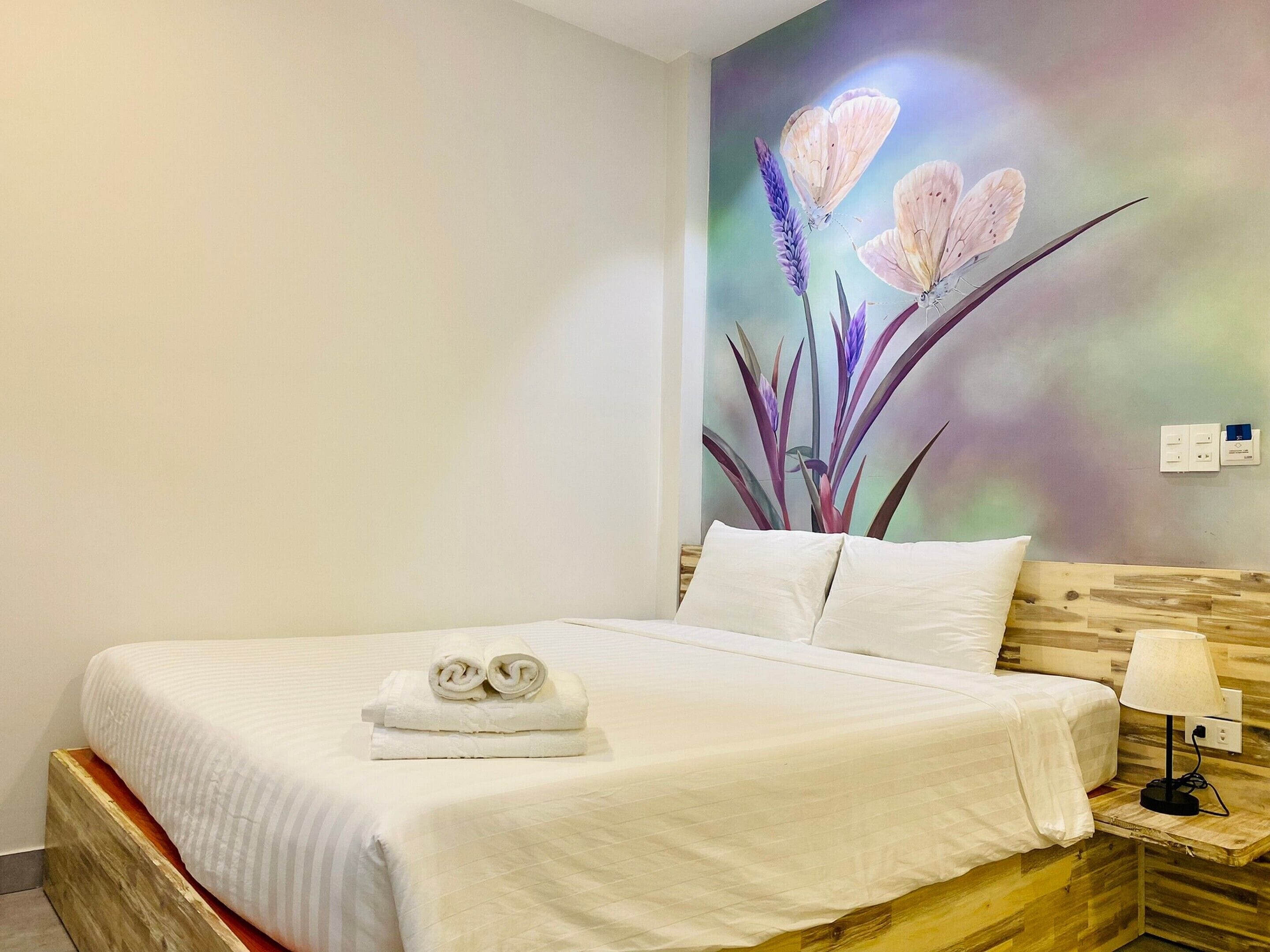 A neatly made bed with white linens and two rolled towels, a wooden headboard, a bedside table with a lamp, and a wall mural of flowers.