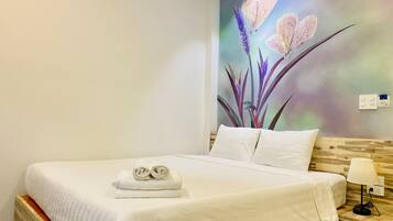 A neatly made bed with white linens and two rolled towels, a wooden headboard, a bedside table with a lamp, and a wall mural of flowers.