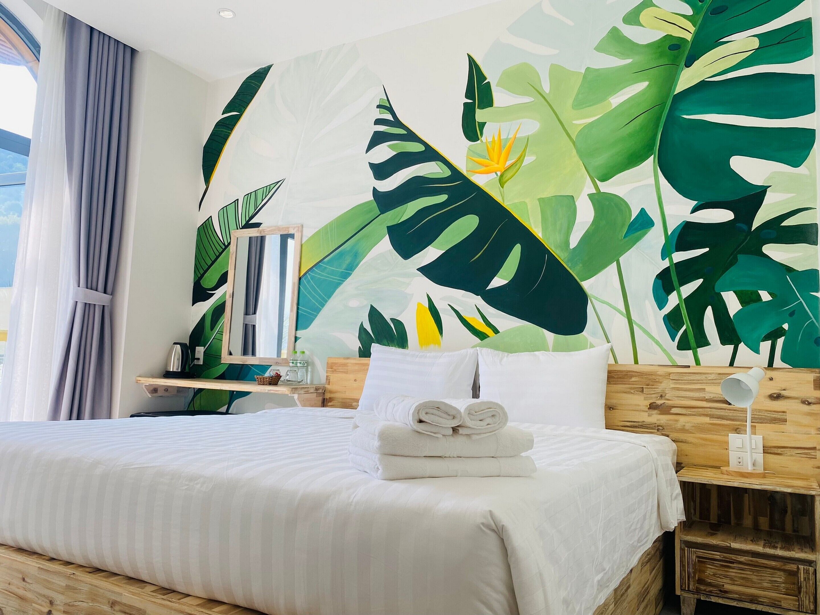 Een hotelkamer met een groot wanddecor van een tropisch blad, een opgemaakt bed met witte lakens en een houten nachtkastje met een lamp.