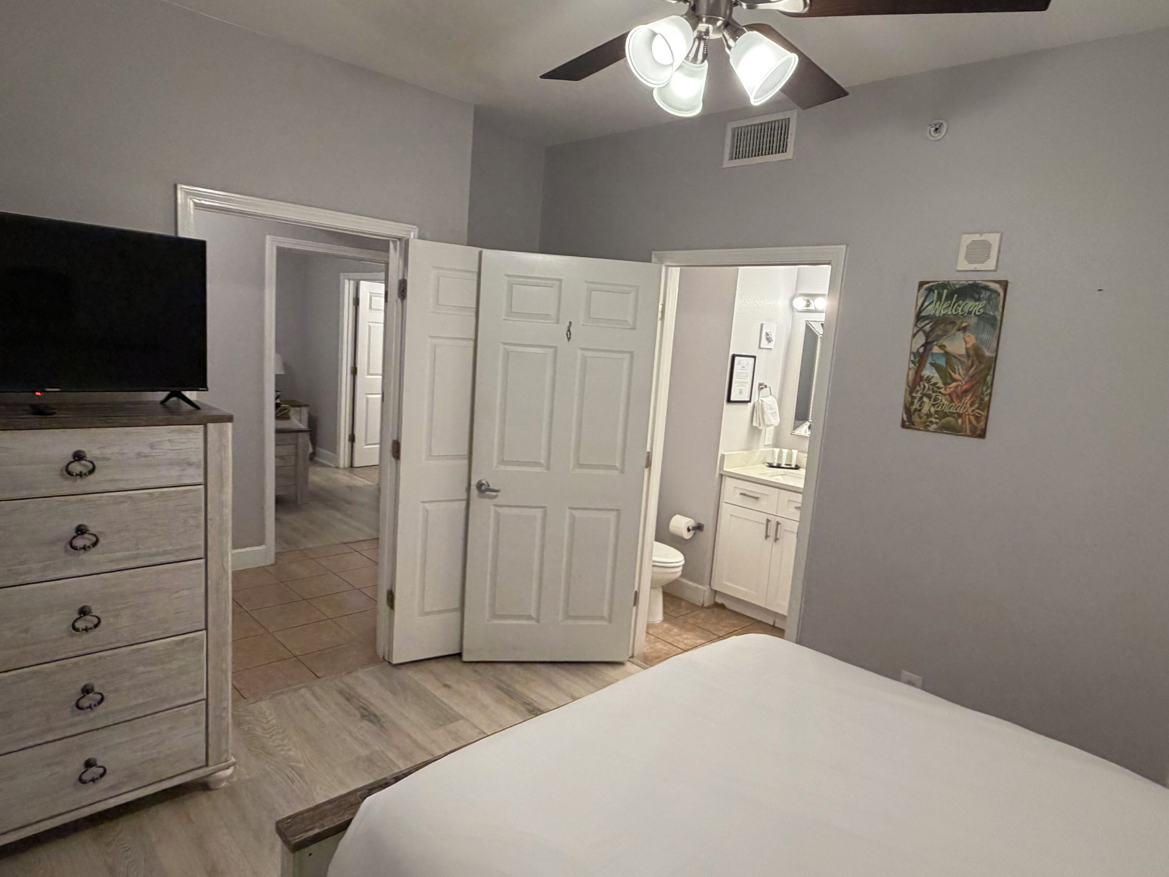 Une chambre à coucher avec un lit, une commode, un ventilateur de plafond et une salle de bain visible à travers une porte ouverte.