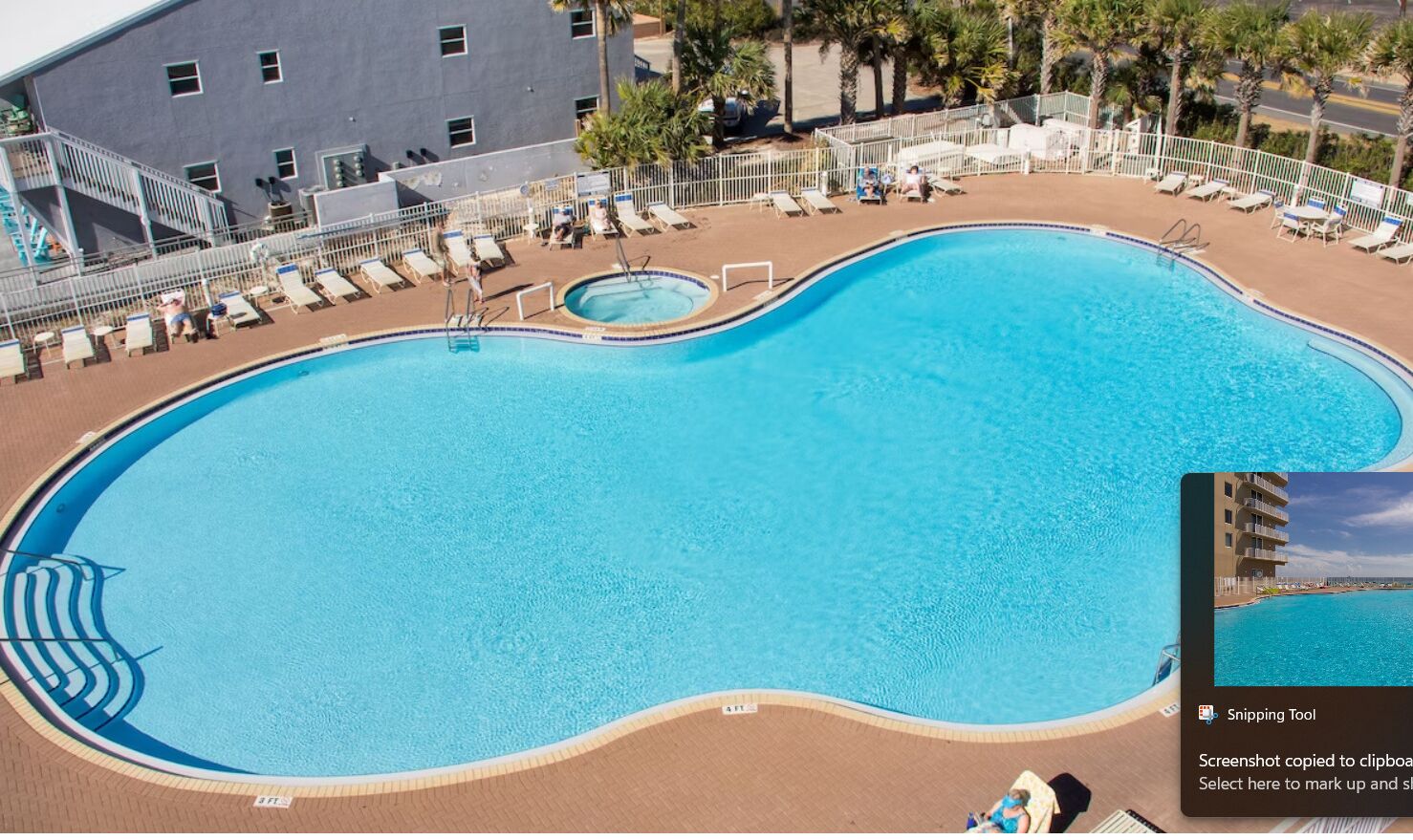 Ein großer, gebogener Swimmingpool mit Liegestühlen und einer kleinen Whirlpool-Anlage, umgeben von einer eingezäunten Fläche und Palmen.