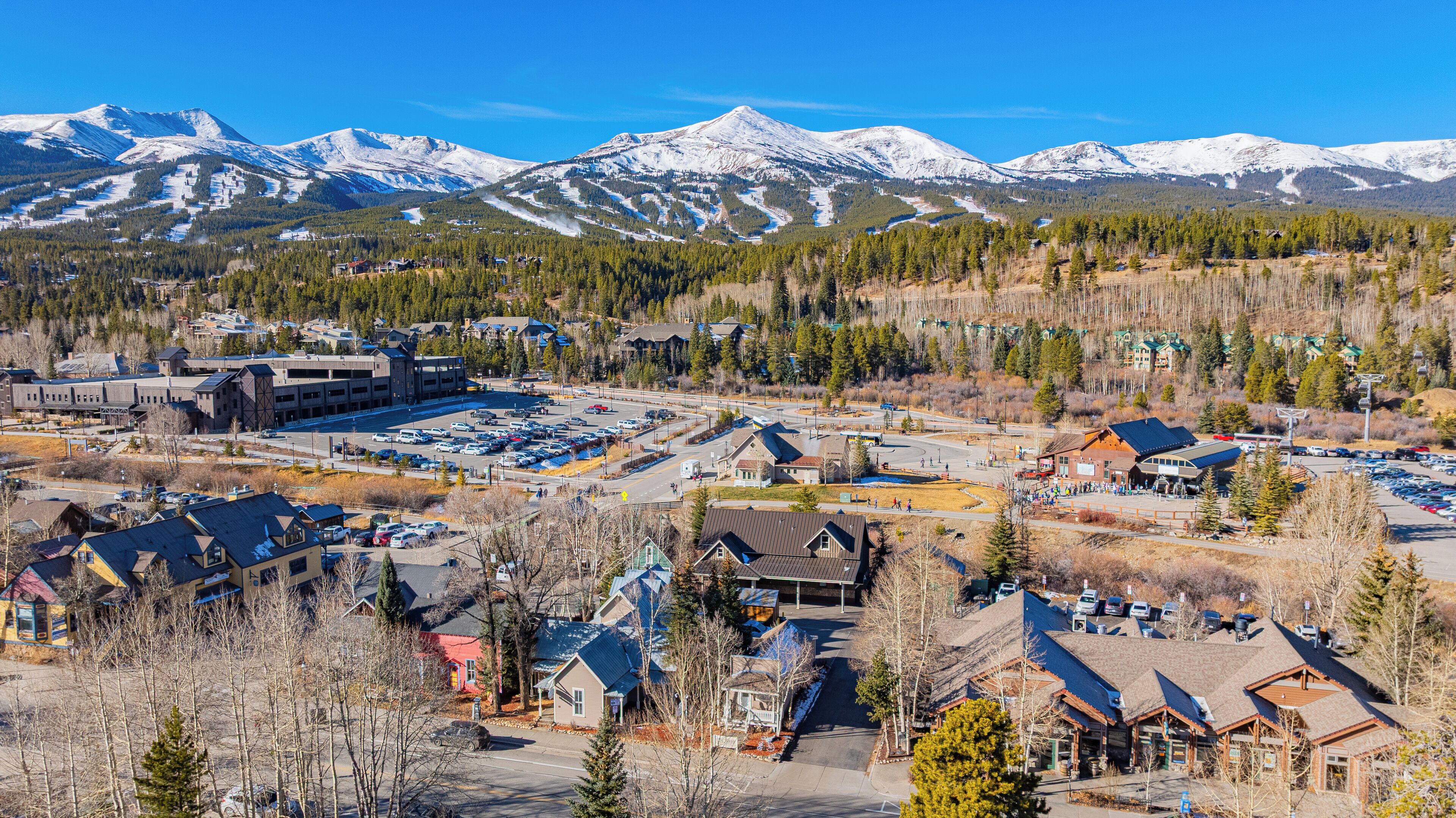 A ski resort with a large parking area, buildings with sloped roofs, and snow-covered mountains in the background.