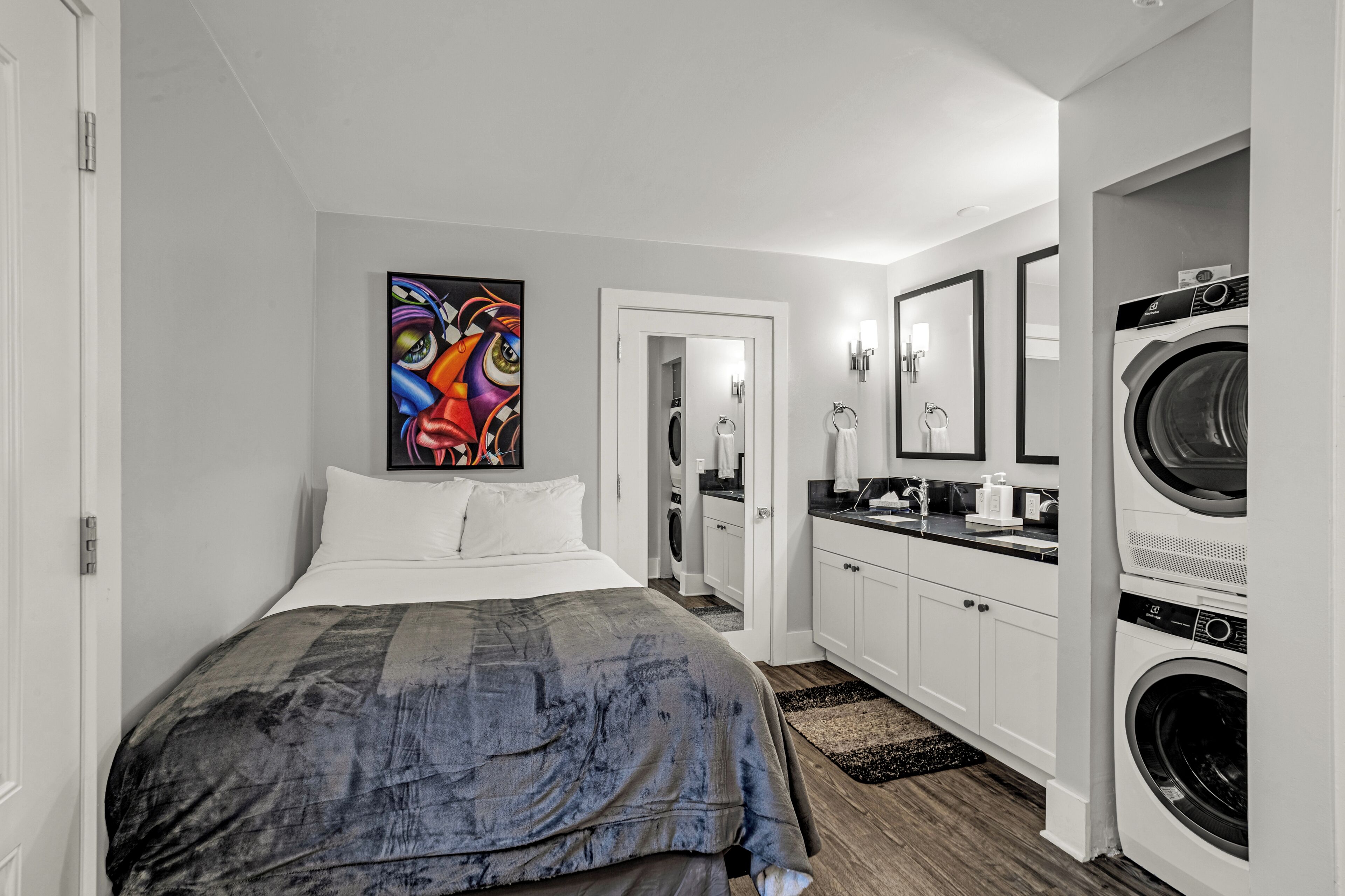 Une chambre à coucher avec un lit, un tableau, une salle de bain avec un miroir et un espace buanderie avec des machines à laver.