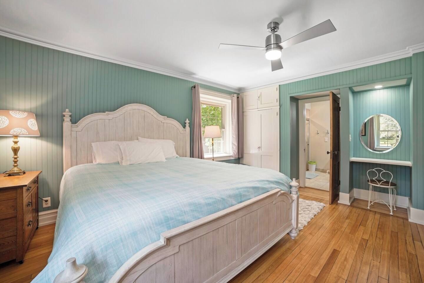 Een slaapkamer met een bed, een plafondventilator, een nachtkastje, een dressoir, een spiegel en een deur die naar een andere kamer leidt.