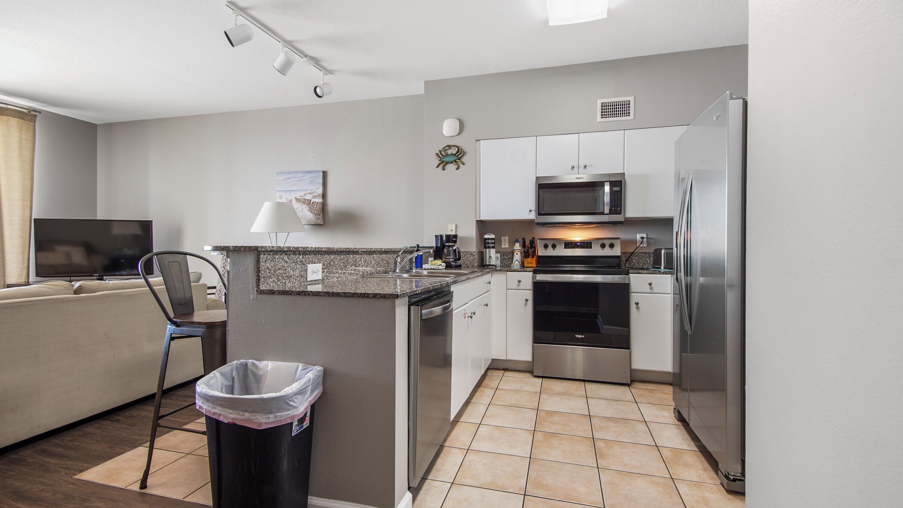 A modern kitchen with white cabinets, a stainless steel refrigerator, a microwave, and an oven. There is a trash can and a chair in the corner.