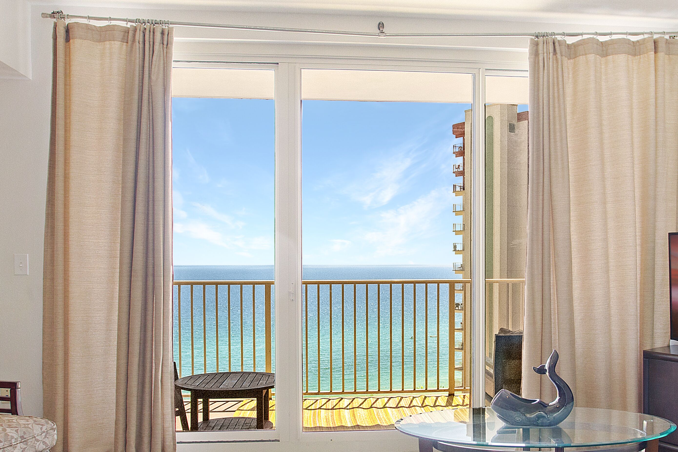 A balcony with a glass table and a decorative item, overlooking a clear blue sea and a building in the distance.