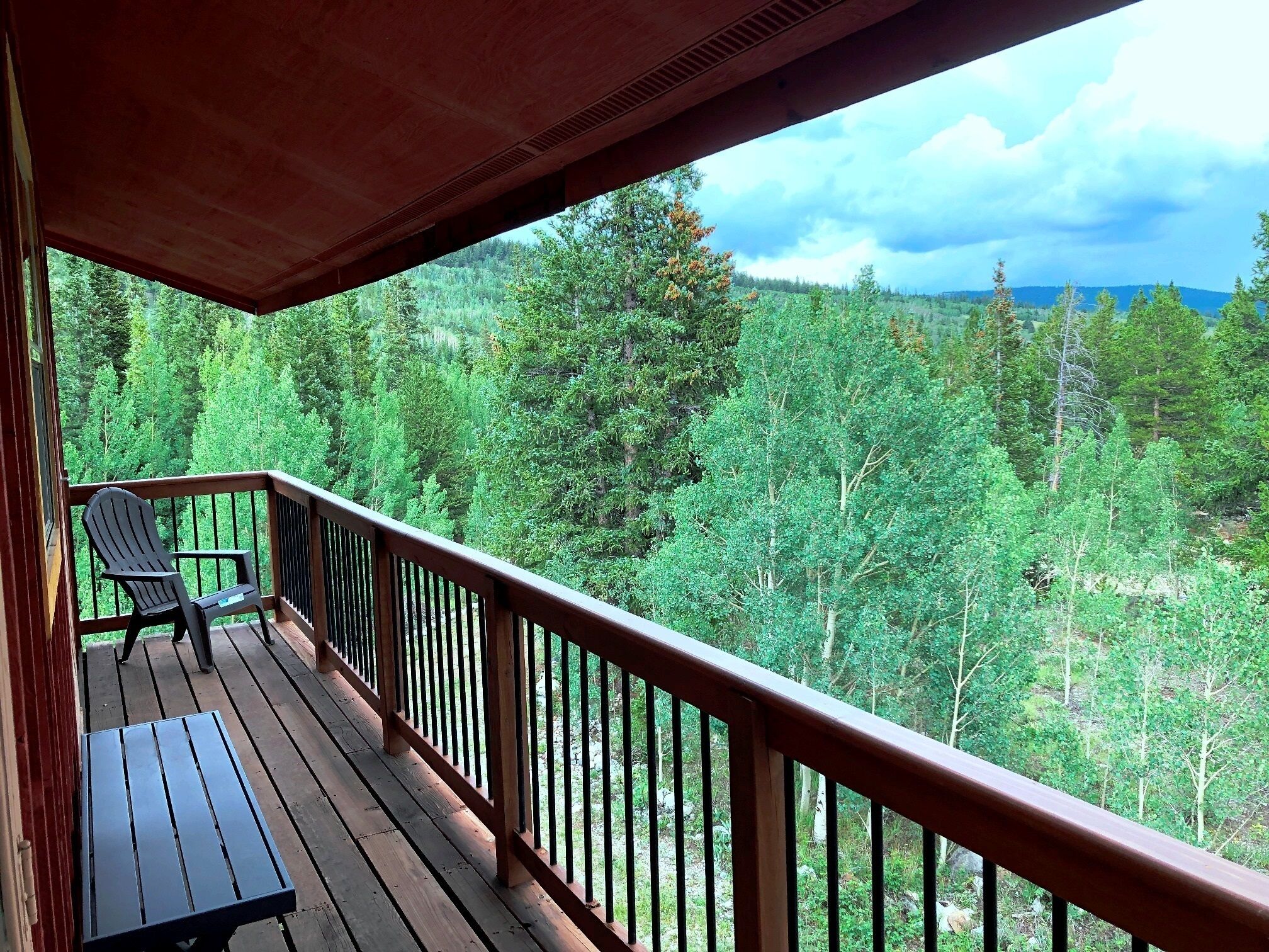 Een houten terras met een bankje en twee Adirondack-stoelen met uitzicht op een bebost landschap.