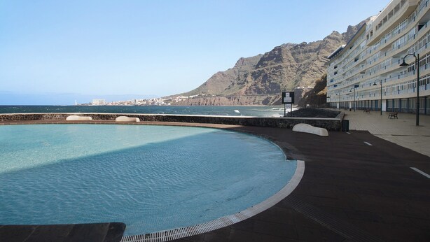 Una piscina con vistas al mar y a las montañas, un edificio con balcones y un camino pavimentado.