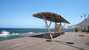 Un paseo marítimo con barandilla de metal, pérgola de madera y un banco.