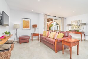 Una sala de estar con un sofá de cuero, mesitas de madera, un televisor montado en la pared y un ventanal con vistas a un patio exterior.
