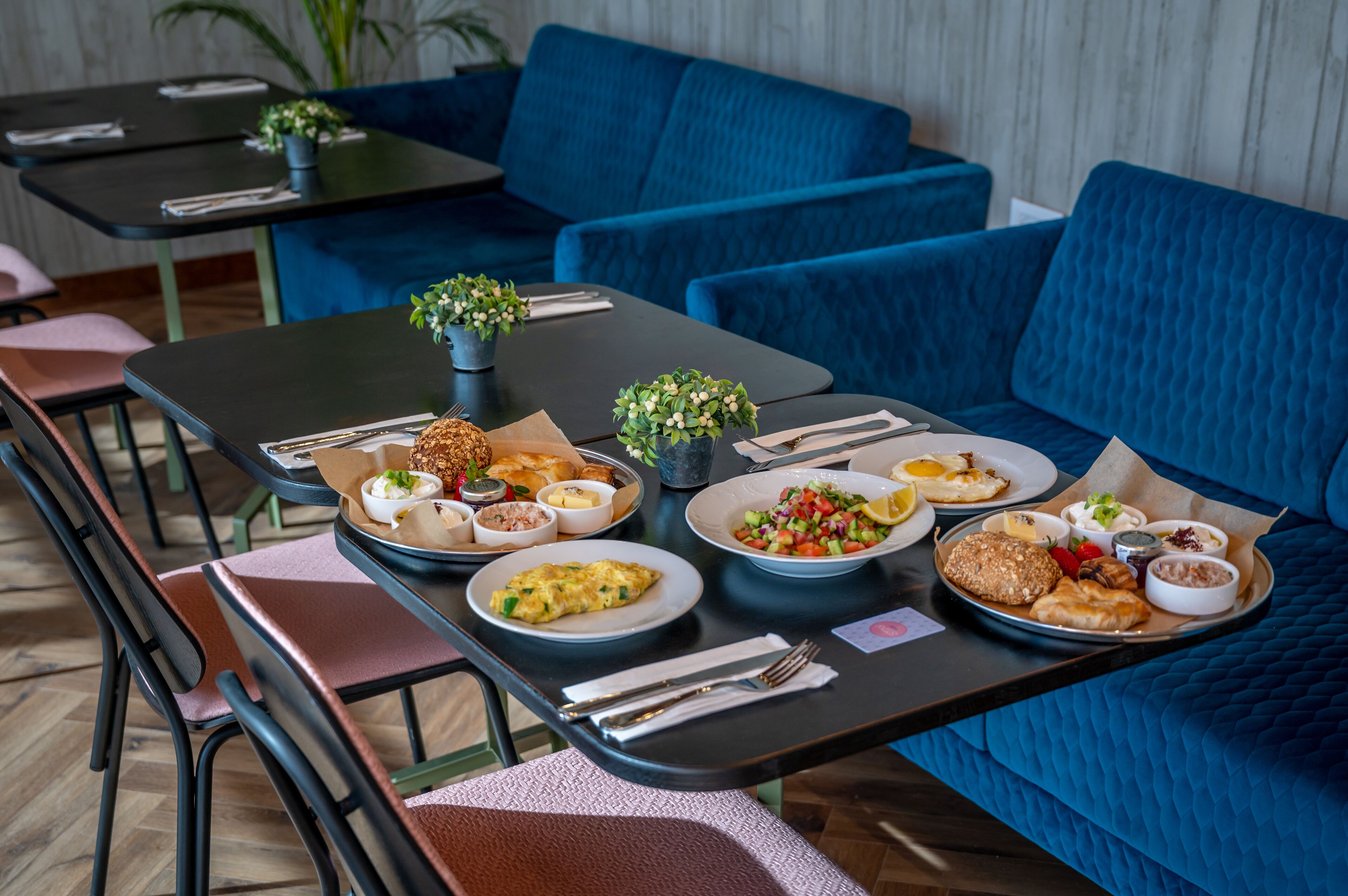 A dining area with tables set for breakfast, including plates of eggs, toast, and salads, in a modern interior with blue upholstered seating.