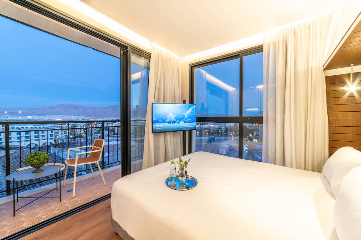 A hotel room with a large bed, a TV on the wall, a balcony with a chair and table, and a view of the city.