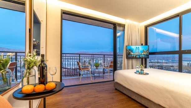 Una habitación de hotel moderna con una cama grande, un balcón con mesa y sillas, un televisor y vistas a la ciudad y las montañas.