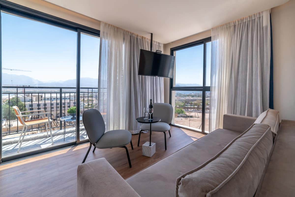 A modern living room with a sofa, two chairs, a small table with a bottle and glasses, a TV, and large windows offering a city view.