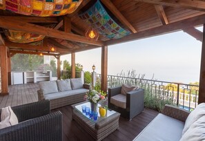 Un espacio de estar al aire libre techado con mobiliario de mimbre, una mesa de centro de vidrio y vista al mar.