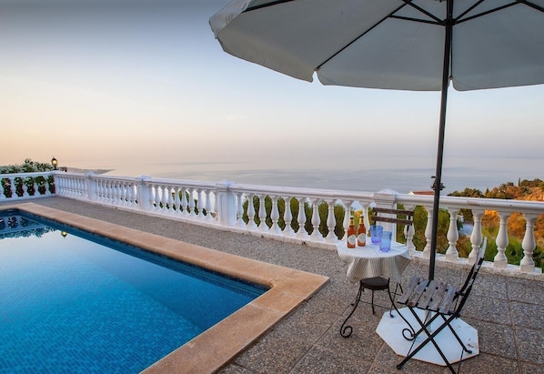 Une piscine bordée d’une balustrade blanche, une table avec des boissons et une vue sur la mer.