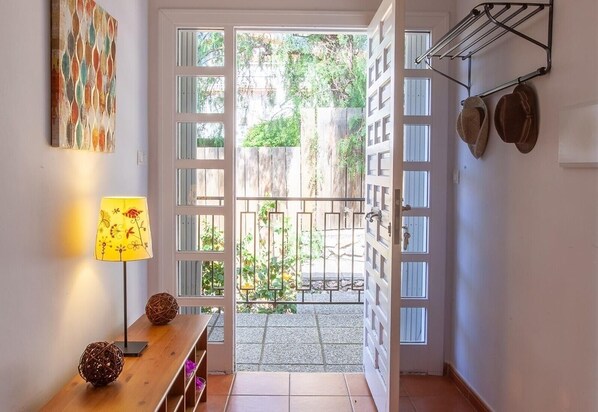 Puerta abierta que da a un patio con una reja metálica, una lámpara, una cesta de mimbre y un perchero con sombreros.