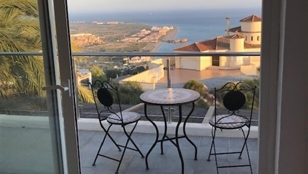 Een balkon met twee stoelen en een tafel, met uitzicht op een kustlandschap met gebouwen en zee.