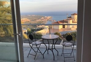 Un balcon avec deux chaises et une table, donnant sur un paysage côtier avec des bâtiments et une vue sur la mer.
