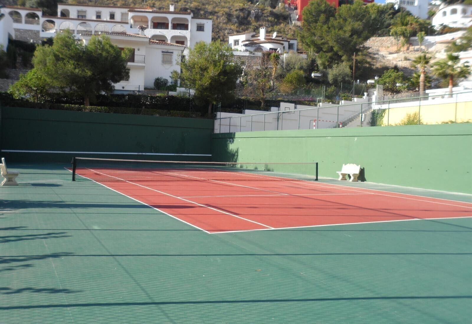 Una cancha de tenis con una red, rodeada de un muro verde y árboles.