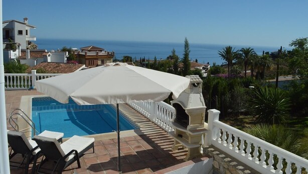 Ein Poolbereich mit weißem Sonnenschirm, Liegestühlen und Blick aufs Meer.
