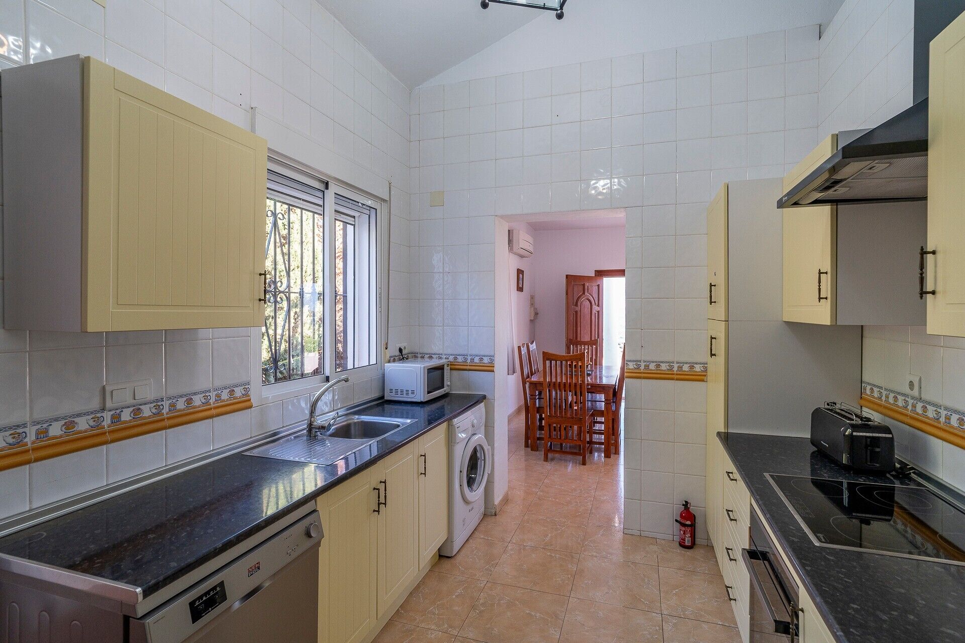 A kitchen with a washing machine, a dining area with chairs, and a tiled floor.