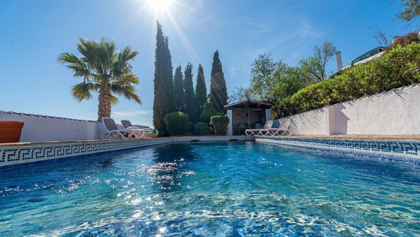 Piscina límpida com espreguiçadeiras e um pequeno edifício, tudo envolto por árvores e um muro branco.