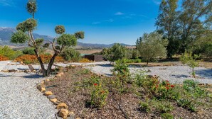 A garden with various plants and trees, a gravel path, and a distant view of mountains. - Villa Las Alondras with Lake and Mountain View, Pool, Garden & Wi-Fi (La Viñuela)