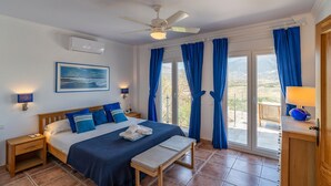 A bedroom with a bed, bedside tables, a ceiling fan, a window with curtains, and a view of the outdoors. - Villa Las Alondras with Lake and Mountain View, Pool, Garden & Wi-Fi (La Viñuela)
