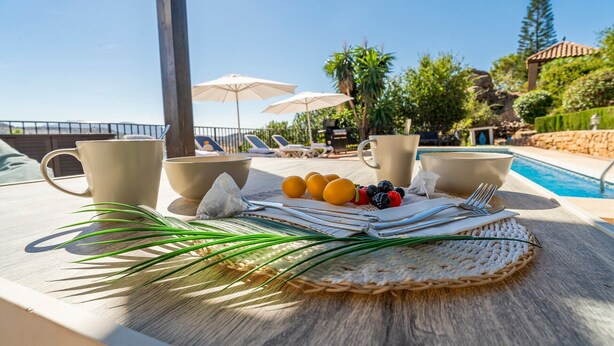 A poolside table with a woven placemat, a cup, a bowl, a napkin, and a fruit arrangement. - Villa Maroma with Mountain View, Pool, Garden & Wi-Fi (La Viñuela)