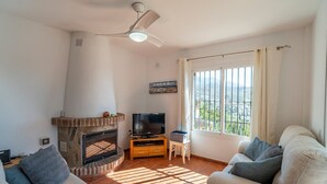 A living room with a stone fireplace, a flat-screen TV, a ceiling fan, and a large window with a view. - Villa Maroma with Mountain View, Pool, Garden & Wi-Fi (La Viñuela)