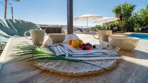 A table with a placemat, cups, cutlery, and fruit, overlooking a pool with umbrellas and lounge chairs. - Villa Maroma with Mountain View, Pool, Garden & Wi-Fi (La Viñuela)