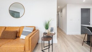 A modern living room with a leather sofa, a round mirror, a small table with a plant, and a kitchen area visible in the background.