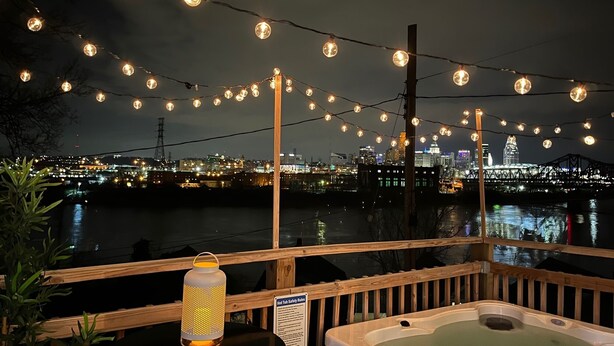 En terrass med ett bubbelbad, ljusslingor och en lykta, med utsikt över en stad och en flod i nattens mörker.