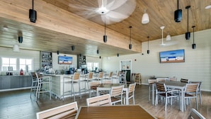Un comedor interior amplio con suelo de madera, varias mesas y sillas, y una zona de bar con pantalla de televisión.