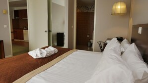 A hotel room with a bed, a wooden headboard, a bedside table with a lamp, and a view of the bathroom and kitchen through an open door.