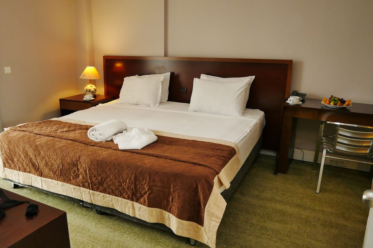 A hotel room with a large bed, a bedside table with a lamp, a chair, and a small table with a fruit bowl.