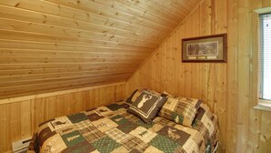 Ein Schlafzimmer mit schrägverdeckter Decke, Holzwänden, einem Bett mit gemustertem Überwurf und einem gerahmten Bild an der Wand.