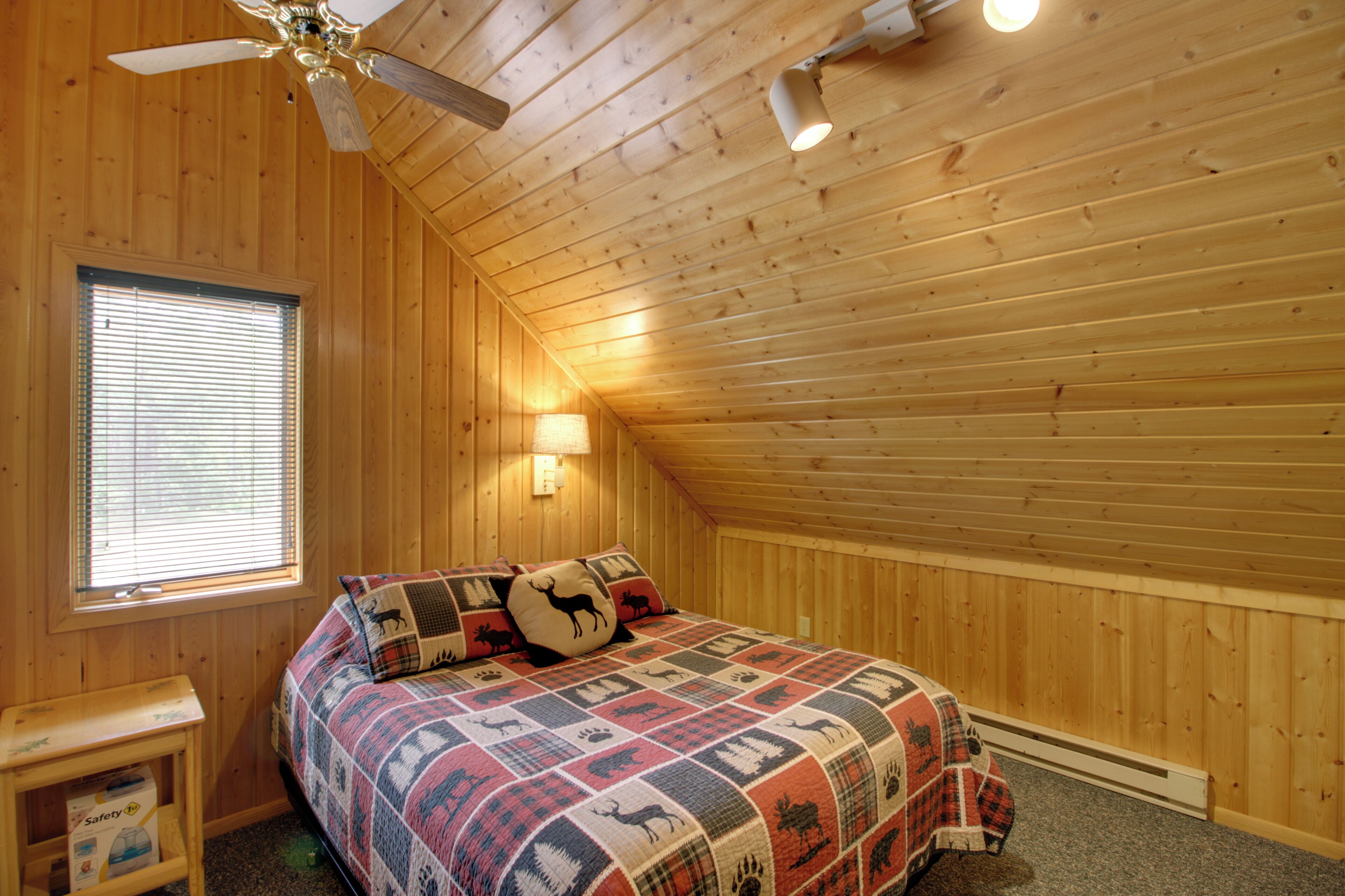 A bedroom with a wooden ceiling, a bed with a patterned quilt, a nightstand, a window with blinds, and a ceiling fan.